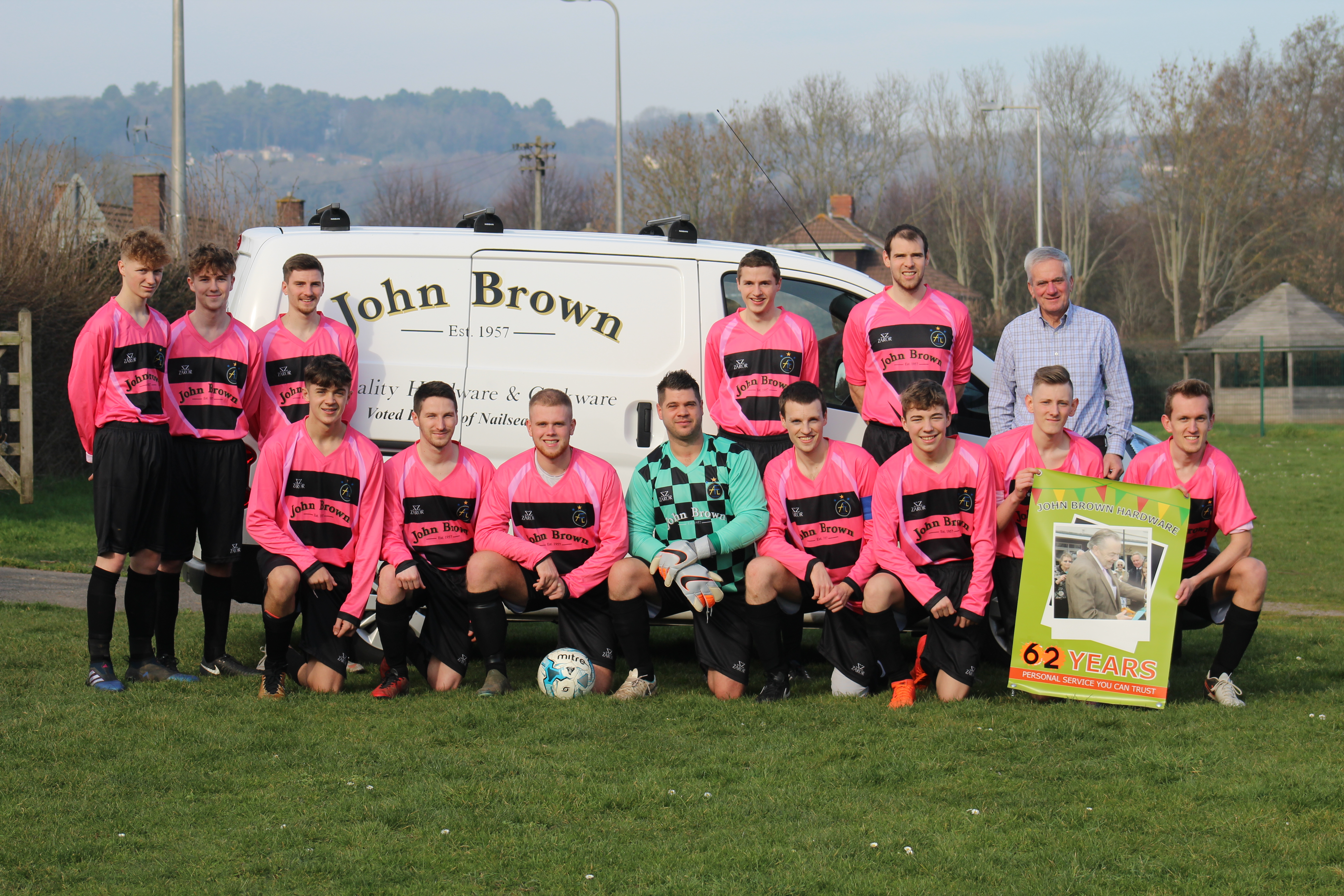 A football team in pink kit lined up in front of a John Brown Hardware van, holding a board reading 62 years