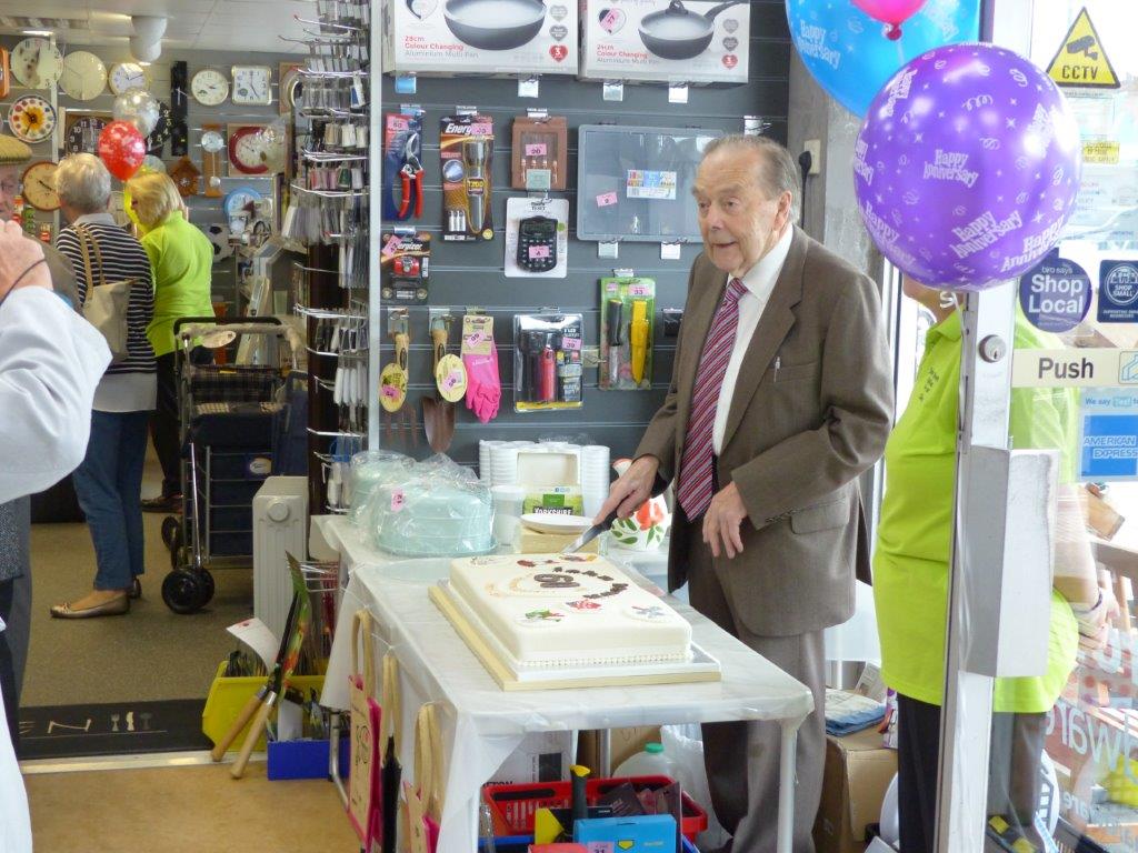 John Brown cutting the shop's 60th anniversary cake beside celebration balloons