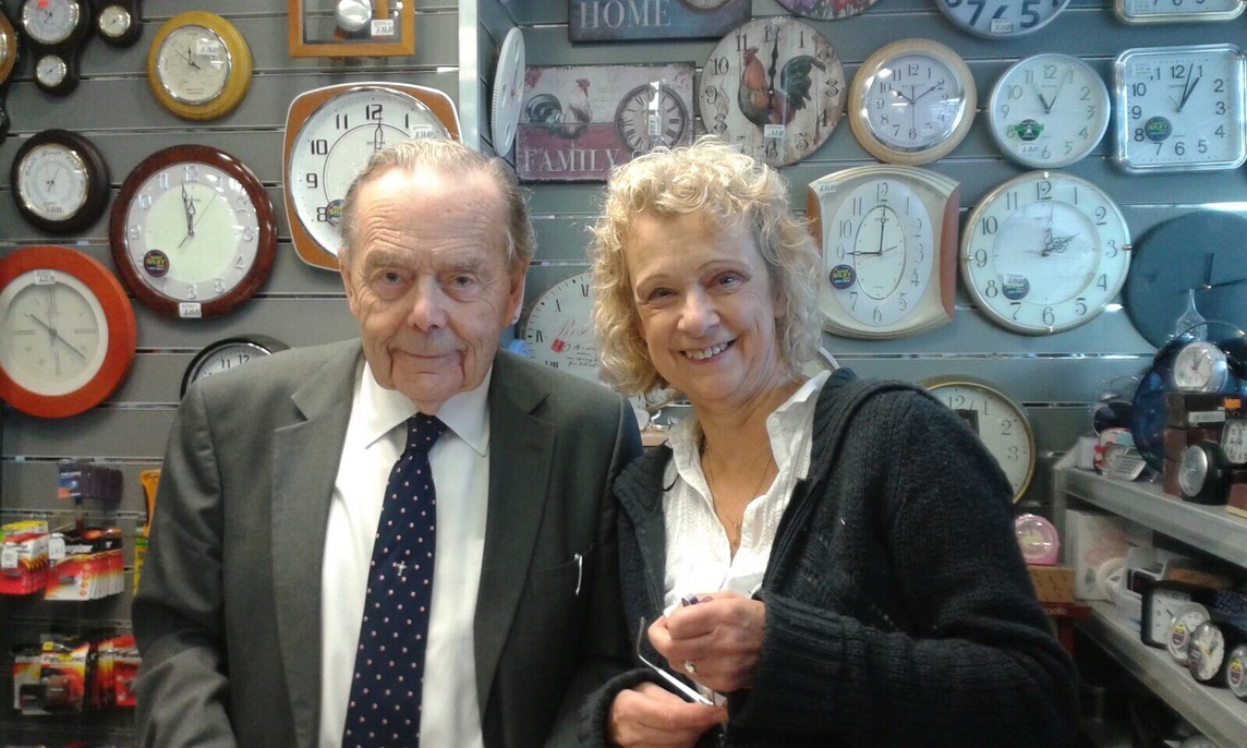 John Brown and his daughter Maggie Fordham standing in front of the shop's wall of clocks